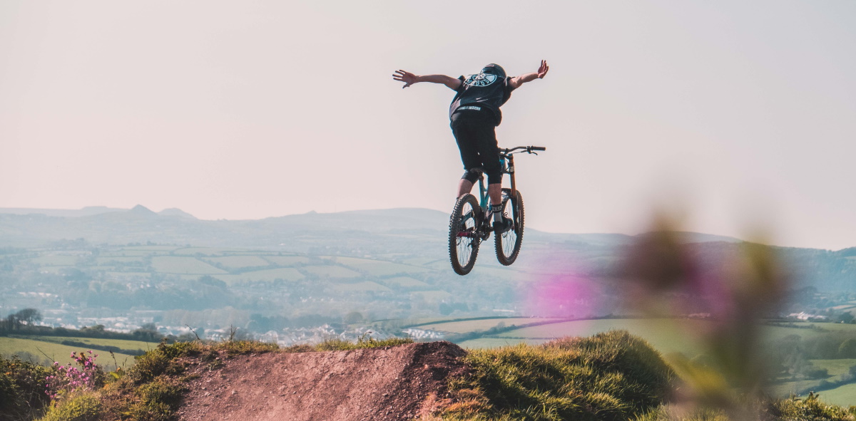 A mountain biker performing a mid-air jump with arms outstretched over a dirt ramp, featuring a scenic rolling countryside background.