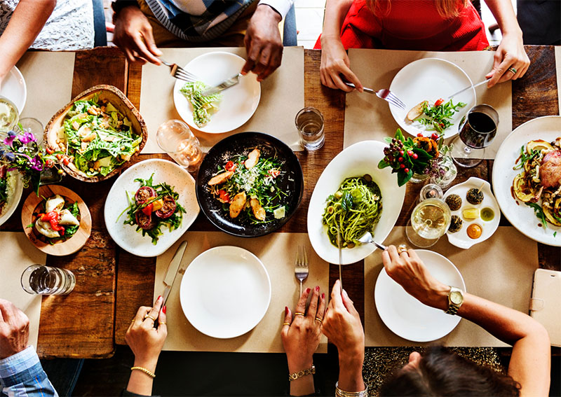 People sitting around a wooden table sharing a colorful meal with salads, pasta, grilled food, and drinks. Hands are reaching for food, creating a lively, communal dining atmosphere.