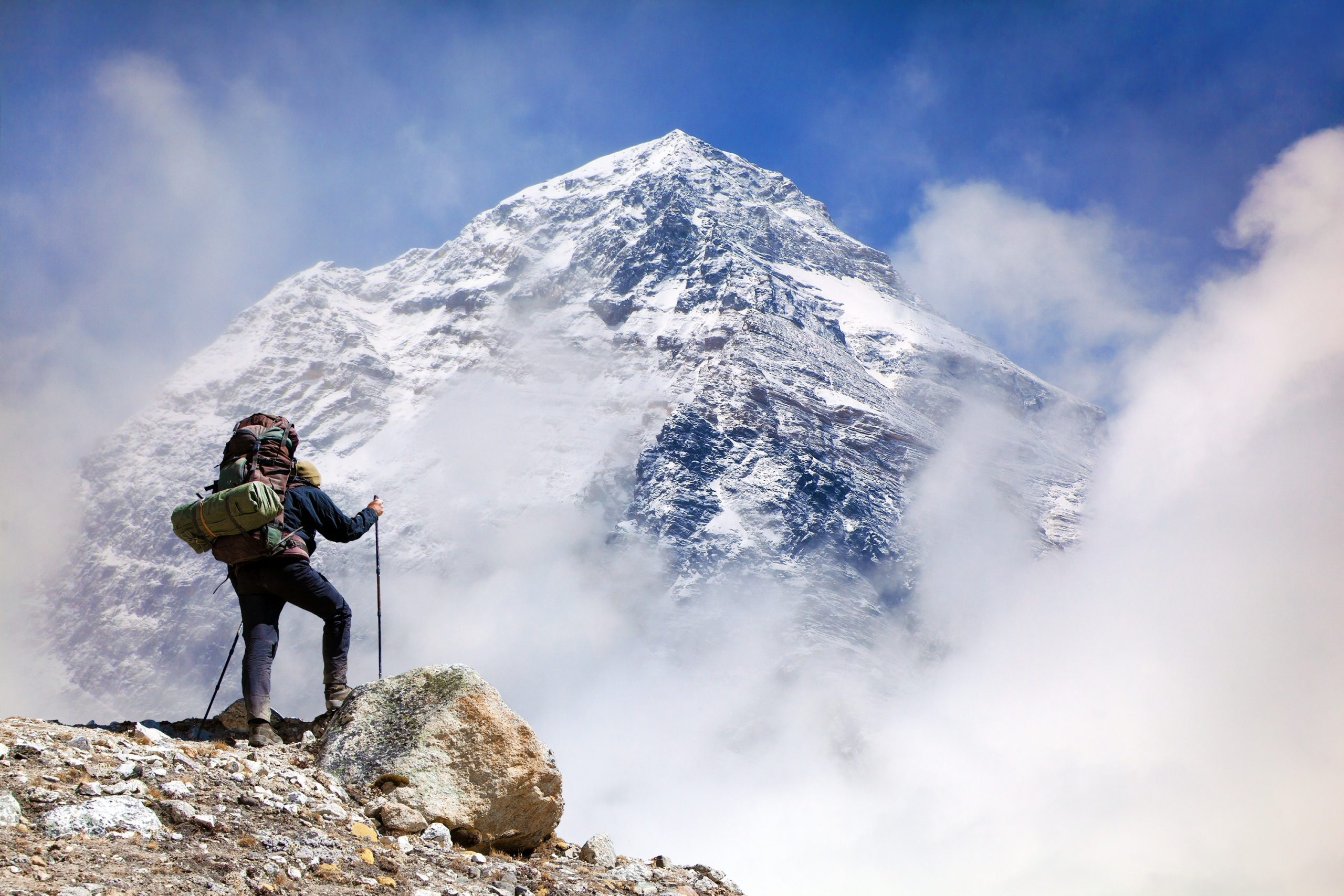 A hiker with a large backpack and trekking poles standing on a rocky ridge, looking toward a massive, snow-covered mountain peak partially obscured by clouds.