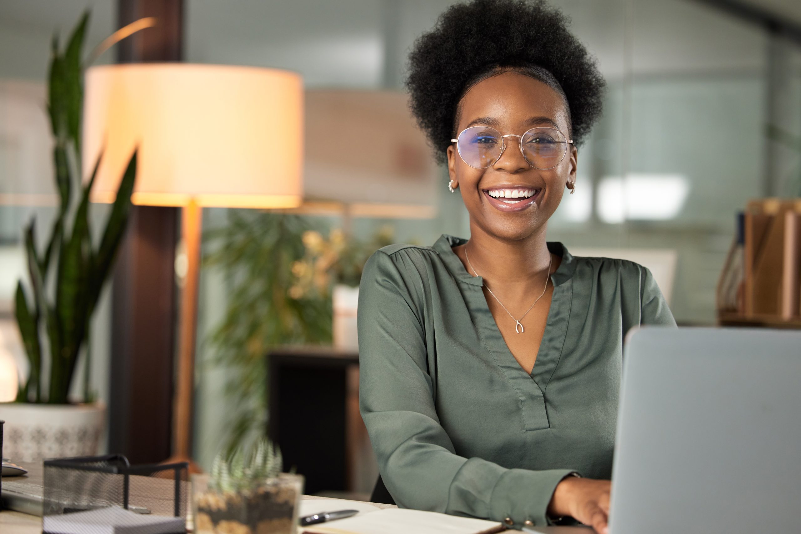 Smiling woman wearing glasses and a green blouse sits at a desk with a laptop in a modern office, with a lamp and plants in the background.
